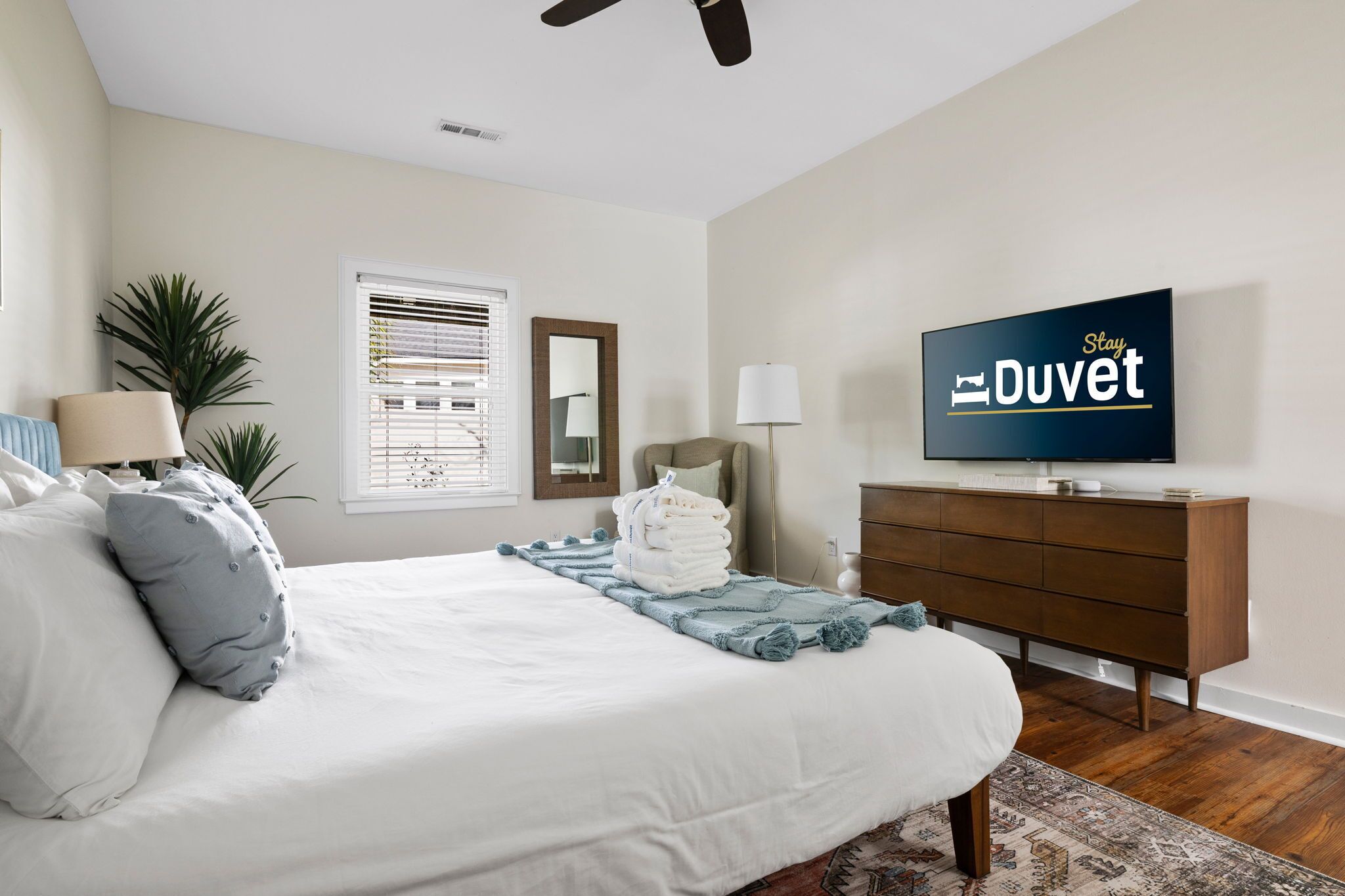 A bedroom with a bed, a TV, a dresser, a mirror, and a window with blinds.