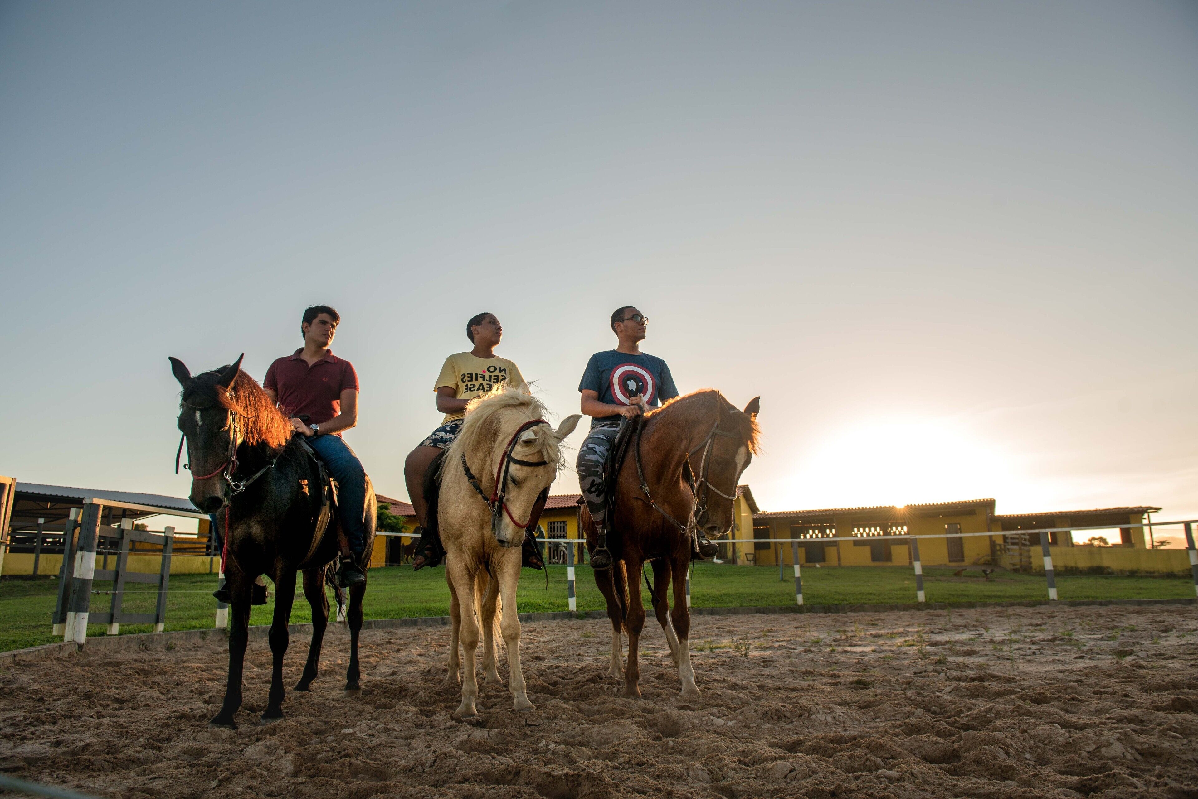Tres personas montando a caballo en un picadero al aire libre durante el atardecer.