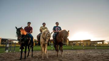 Tres personas montando a caballo en un picadero al aire libre durante el atardecer.