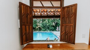 A wooden door leading to a swimming pool with a view of greenery and people in the pool. - Hotel Fazenda Mirage (Conceição de Jacuípe)