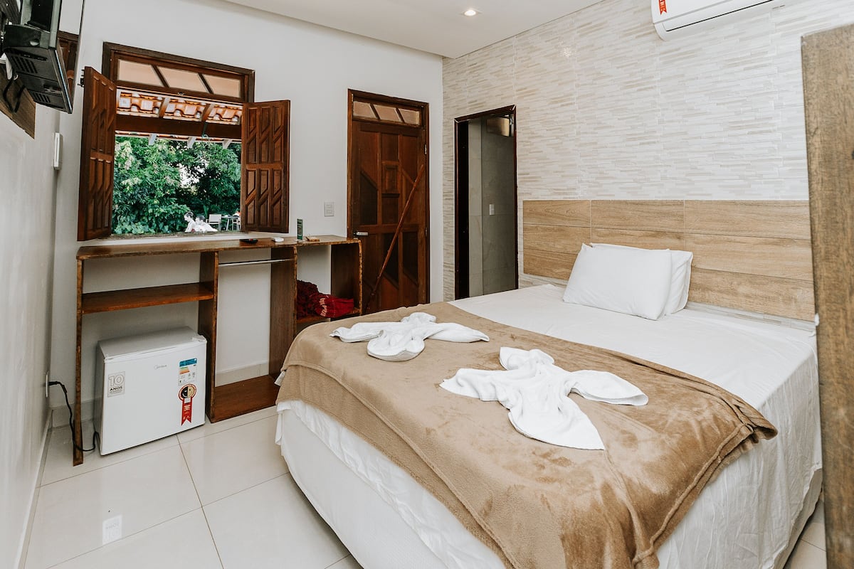 Una habitación de hotel con cama, un escritorio de madera, un refrigerador pequeño y una ventana con vistas a zonas verdes.