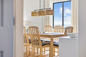 Un comedor con una mesa y sillas de madera, una lámpara colgante de fibras naturales y vistas a través de un amplio ventanal.