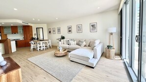 Una sala de estar moderna con sofá, mesa de centro y comedor. Se ve una cocina al fondo, y la habitación tiene ventanas amplias.