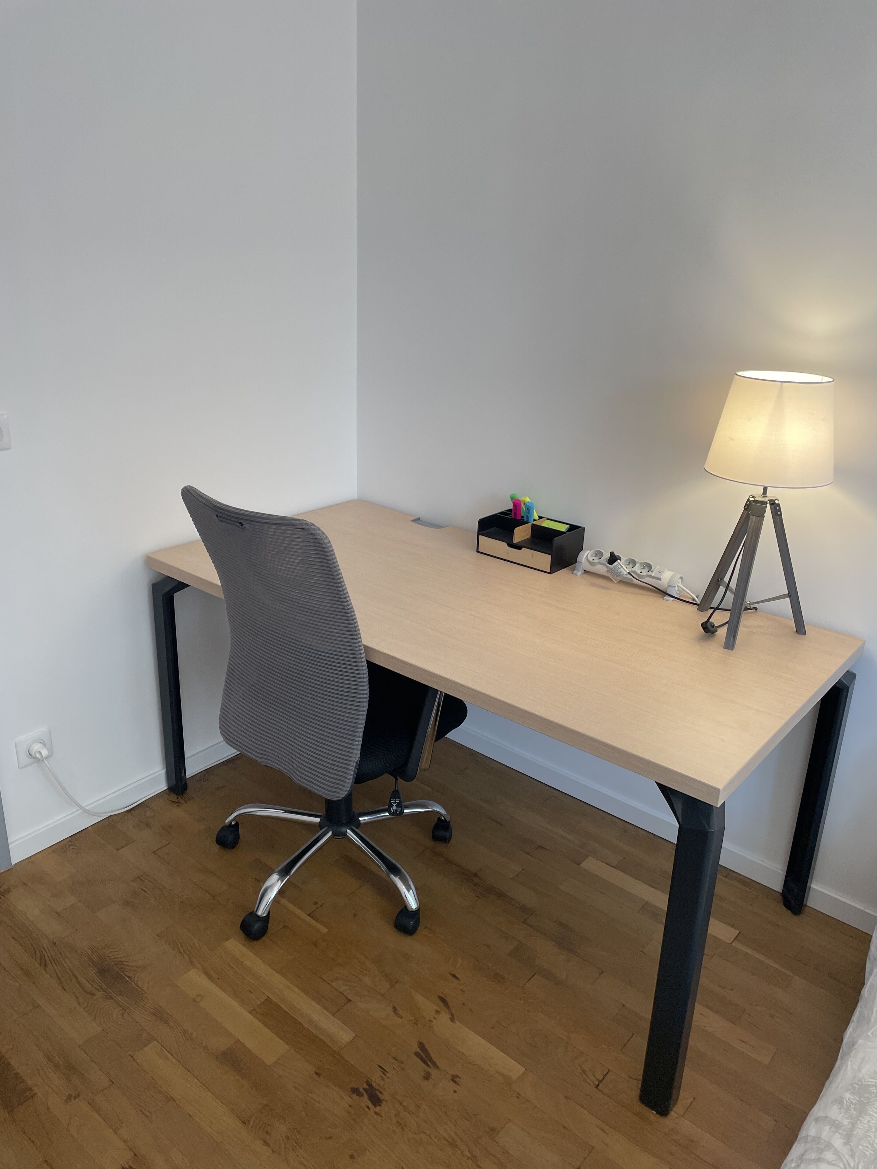 Ein kleiner, aufgeräumter Arbeitsplatz mit einem Holztisch, einem grauen Bürostuhl, einer Lampe und einer Mehrfachsteckdose.