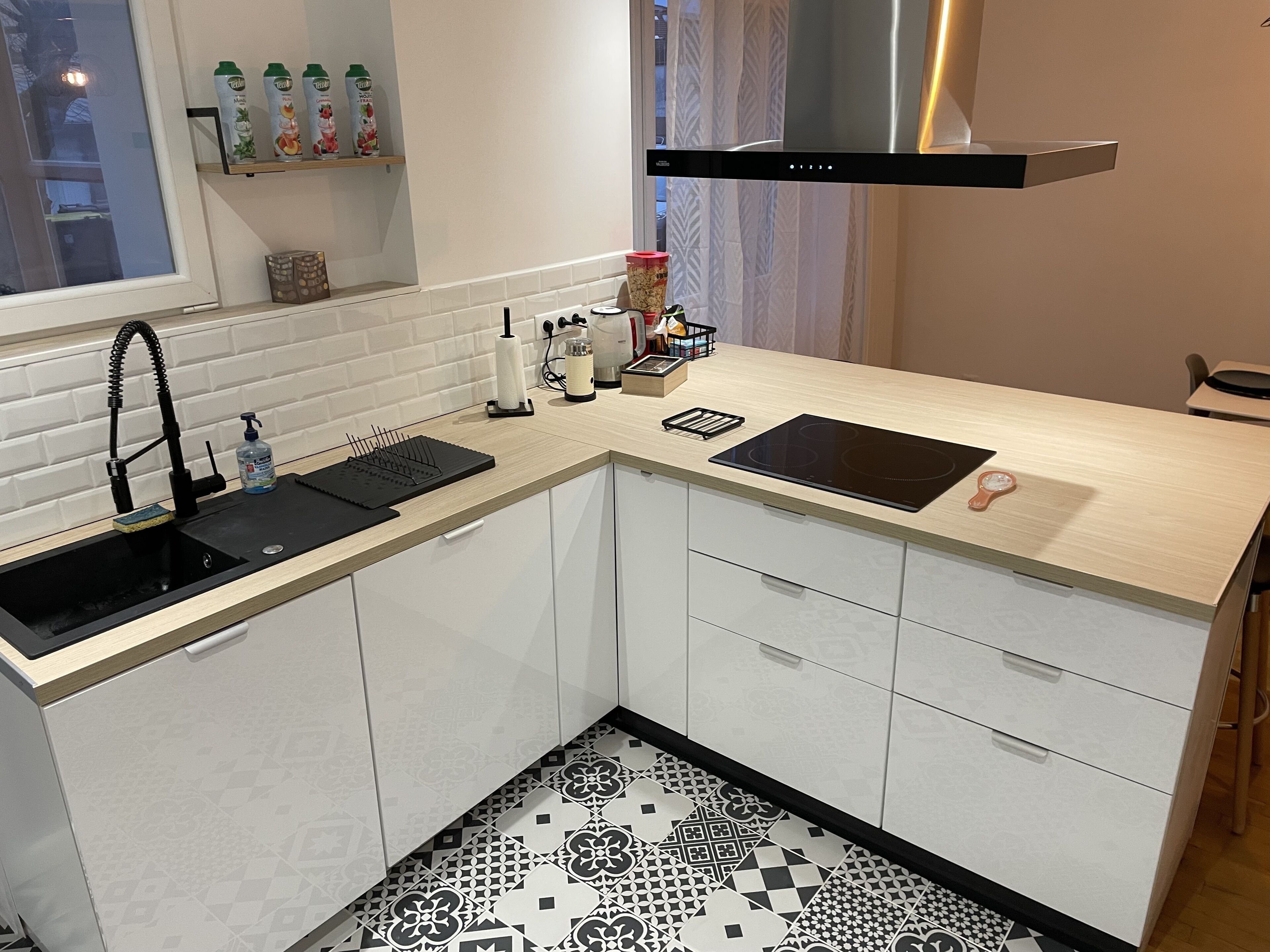 Une cuisine de taille réduite avec un évier noir, des armoires blanches, un pare-feu décoré et un sol carrelé en noir et blanc.