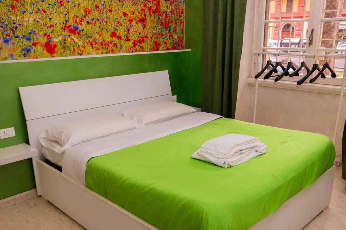 Ein Bett mit grüner Bettdecke und weißen Kissen, ein weißer Nachttisch, ein Fenster mit Blick auf Gebäude und ein farbenfrohes Blumenbild an der Wand.