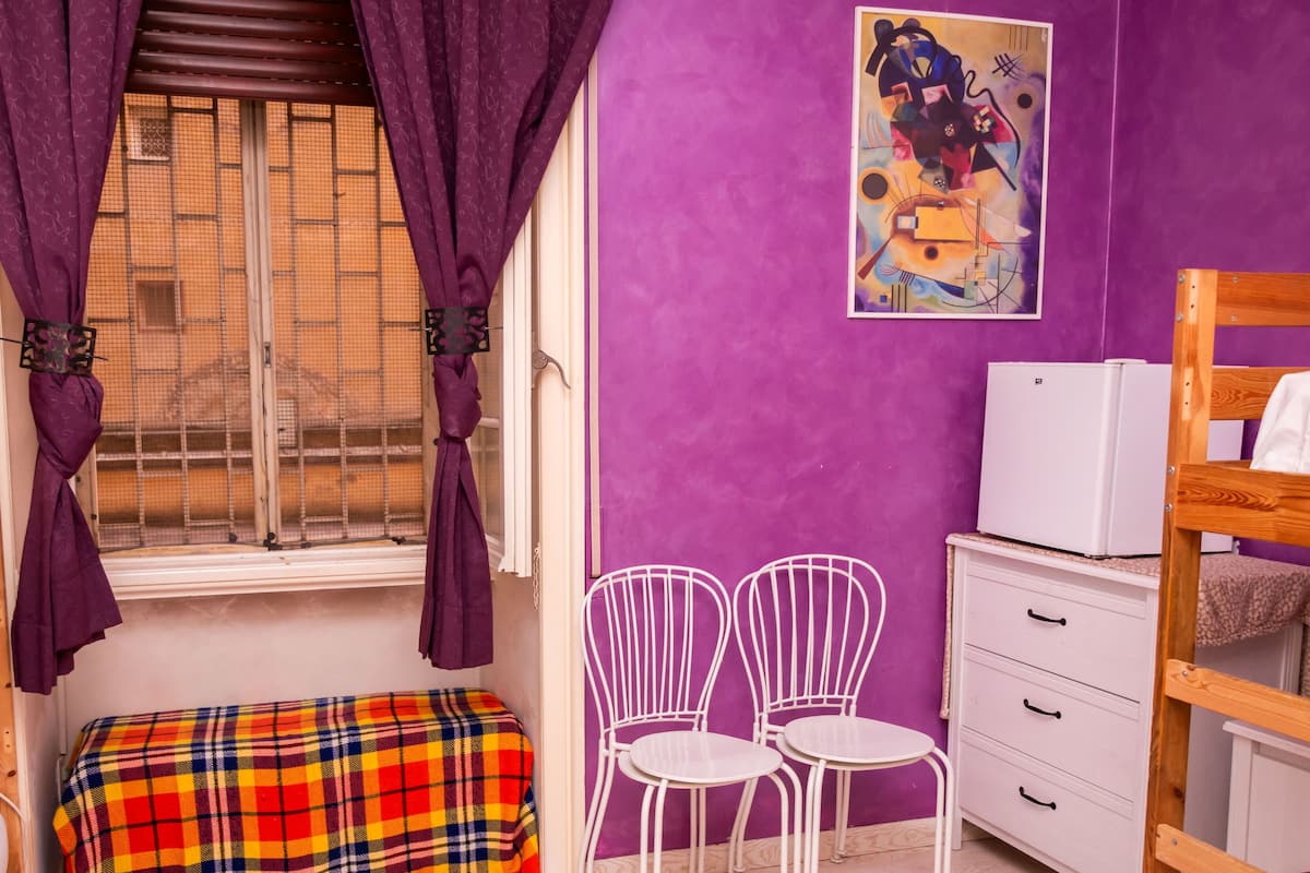 Ein Zimmer mit Etagenbetten, einem kleinen Tisch, zwei Stühlen, einer bunten Decke und einem Fenster mit violetten Vorhängen.