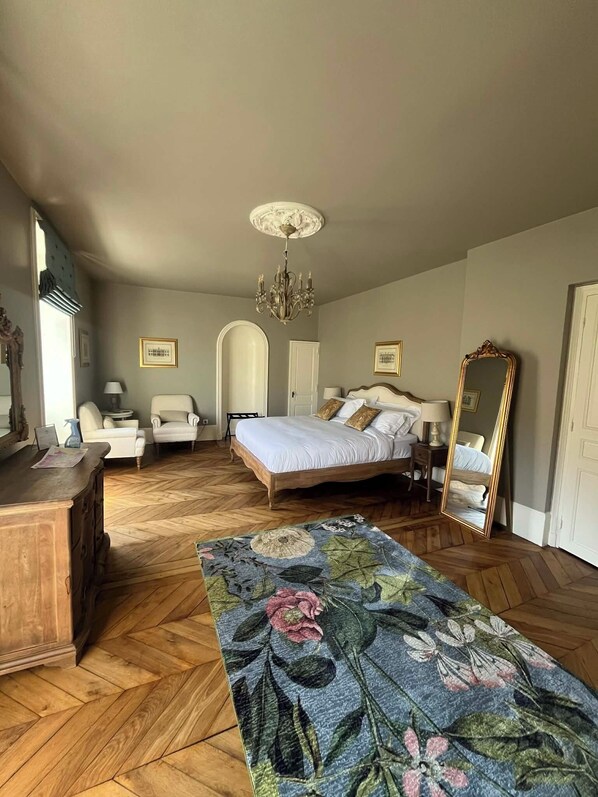 Una camera da letto con pavimento in legno a spina di pesce, tappeto decorato con motivi floreali, un letto con lenzuola bianche, un lampadario, uno specchio grande e un comò in legno.