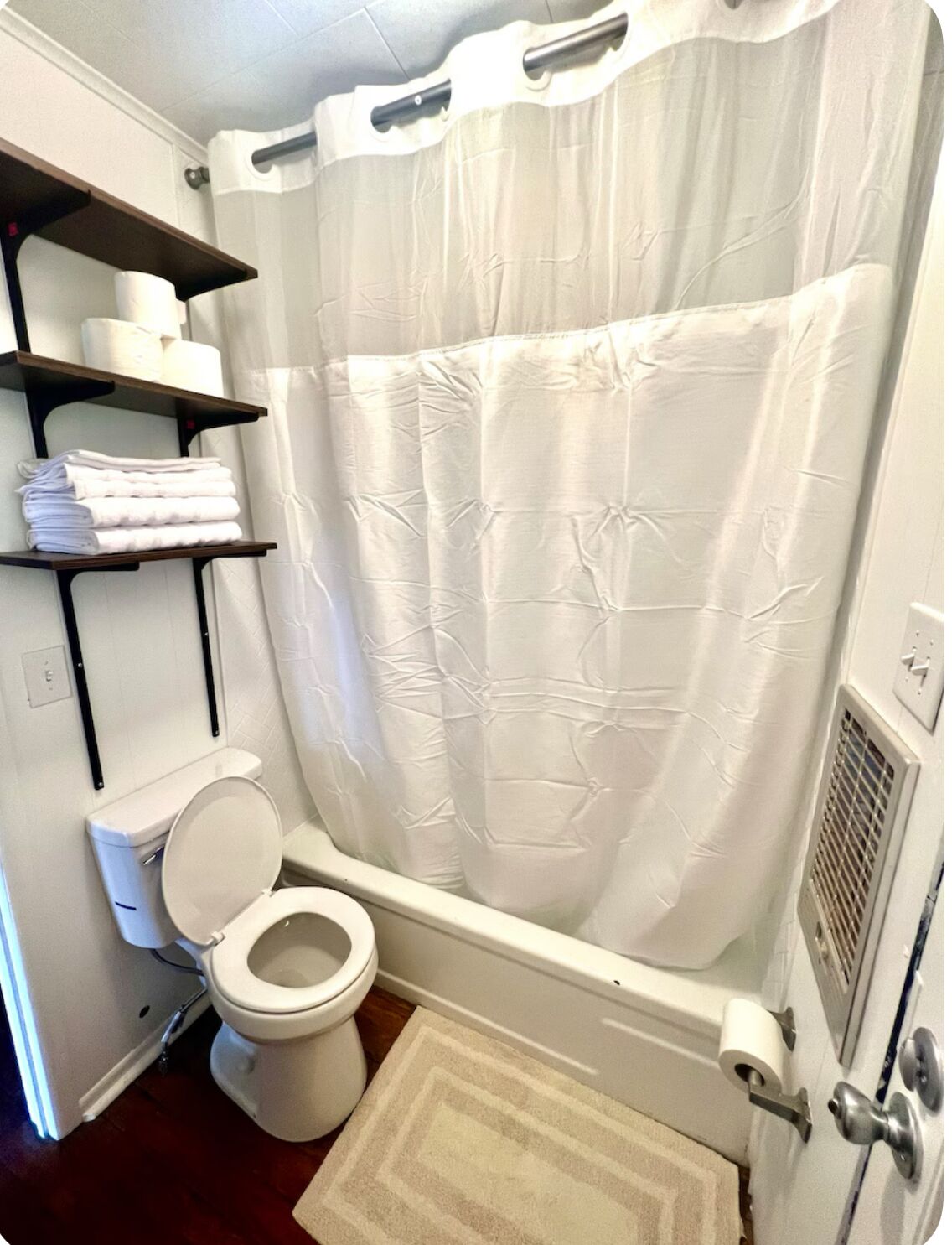 Et badeværelse med toilet, badeforhæng, hylde med håndklæder og en lille måtte.