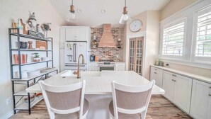Una cocina con isla central, armarios blancos, pared decorada con ladrillo e iluminación colgante.