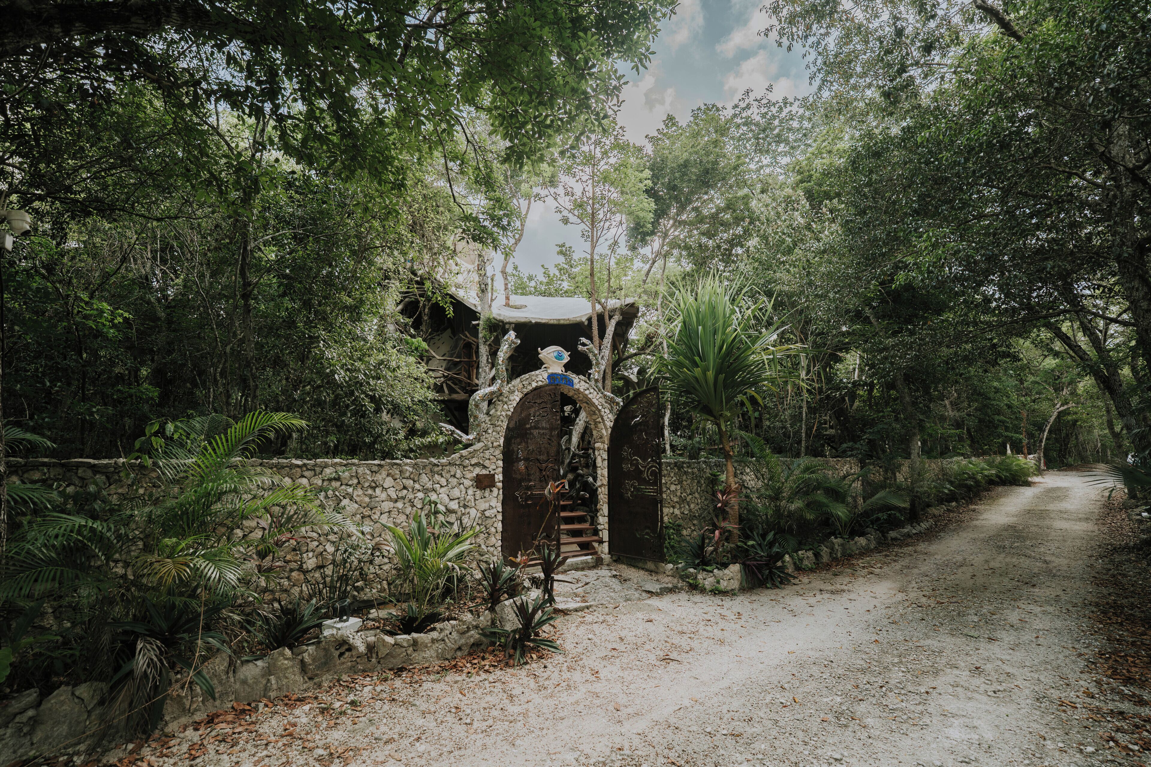 A rustic stone pathway leading to a wooden gate with a decorative archway, surrounded by lush greenery.