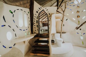 Habitación con un diseño arquitectónico único que presenta una escalera hacia la zona de la cama, un inodoro blanco y un techo con ramas y botellas colgantes.
