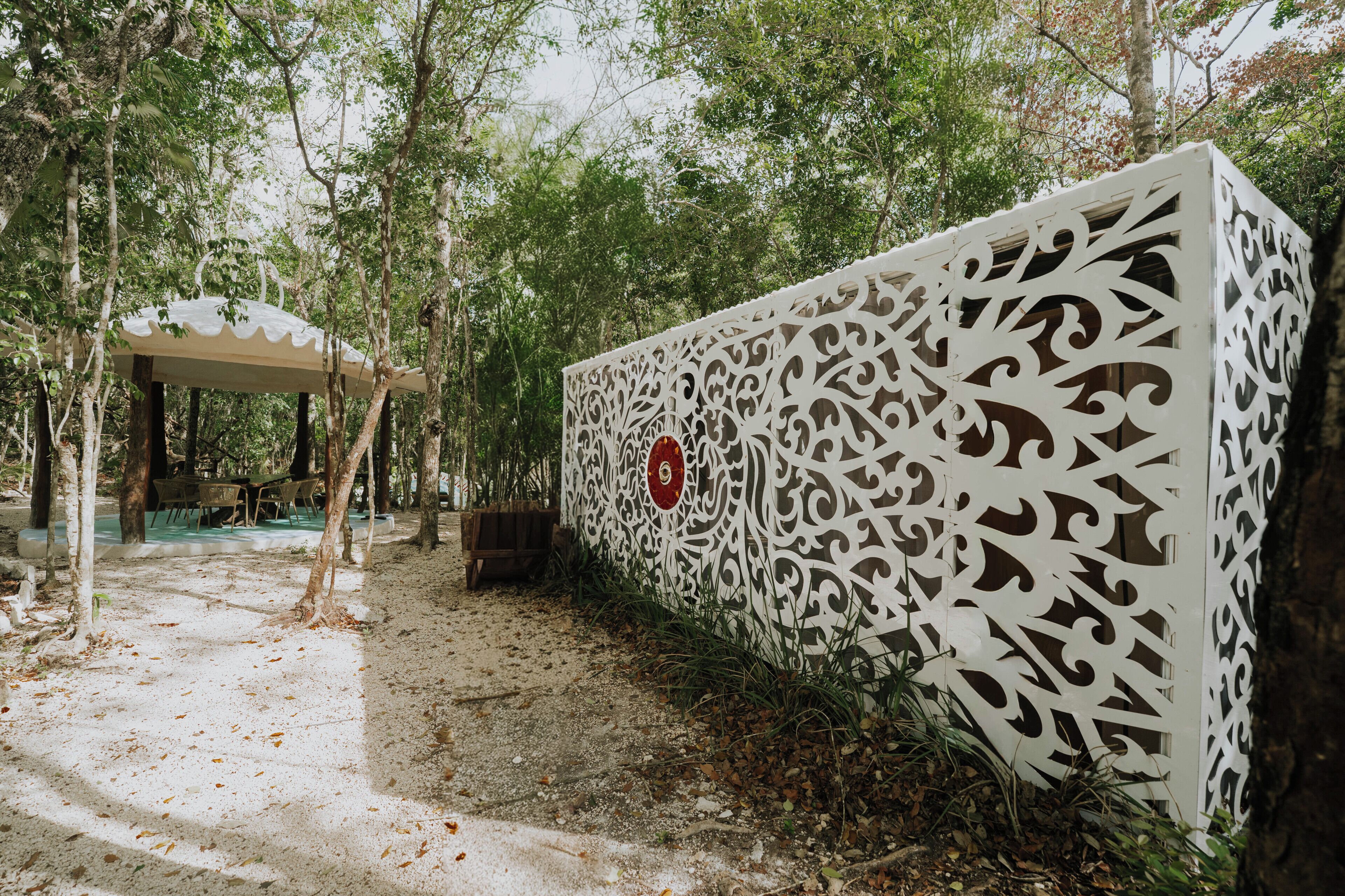 A white decorative wall with intricate patterns, a covered seating area, and a natural forested background.