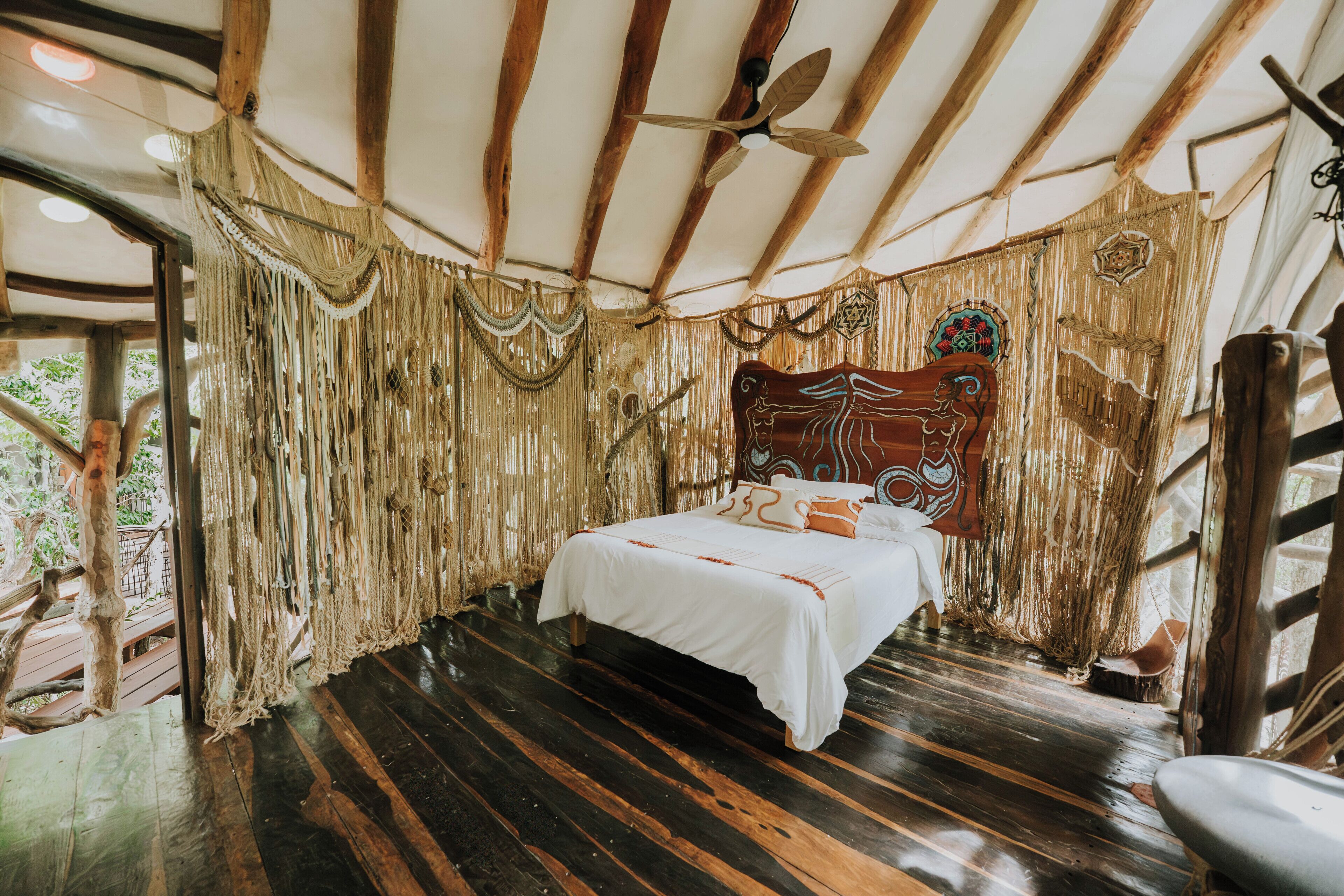 Una habitación con cama, vigas de madera, ventilador de techo y vista a follaje exterior.