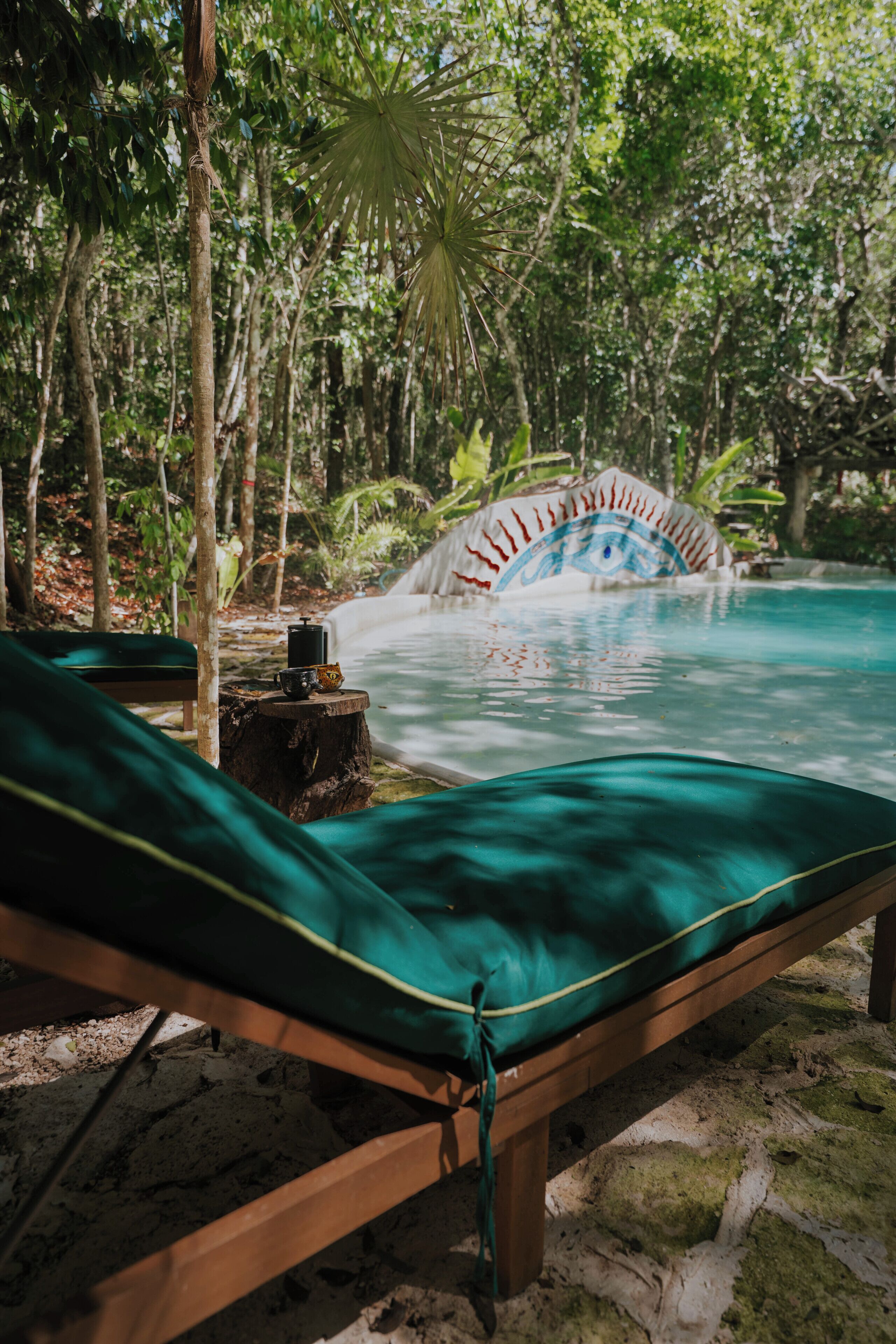 Uma poltrona de madeira com capa verde, uma mesinha com uma chávena e uma tigela, e uma ponte decorativa sobre uma piscina.