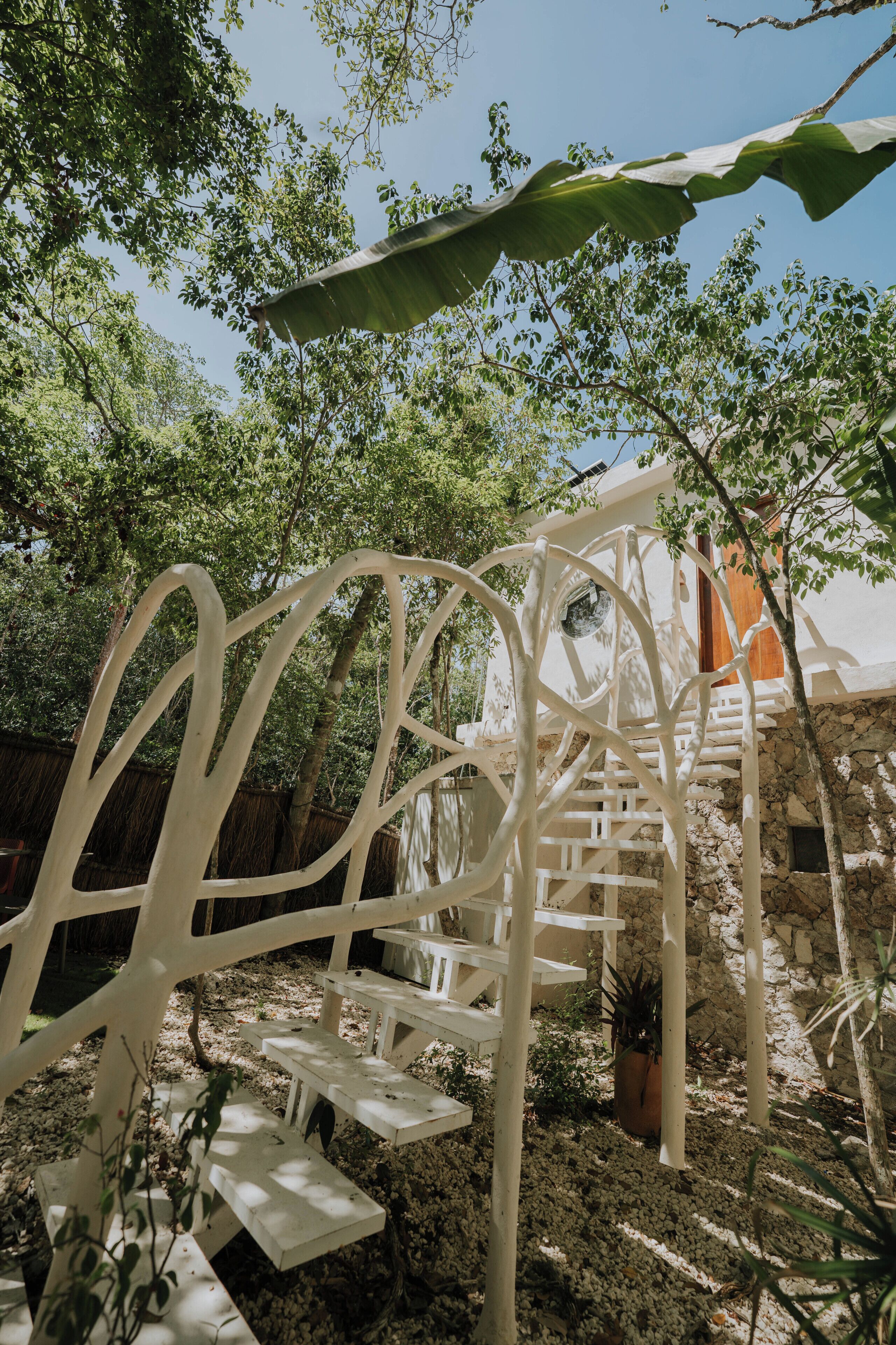 Une structure métallique blanche, dotée de bancs, entourée d’arbres et d’un mur de pierre.