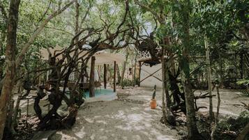 A rustic outdoor seating area with a wooden table and chairs, surrounded by trees and a decorative white structure.