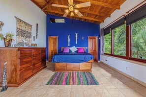 Ein Schlafzimmer mit Bett, Holz-Kopfteil, Deckenventilator, Fenster mit Blick auf Grünflächen und einer Wanddekoration.