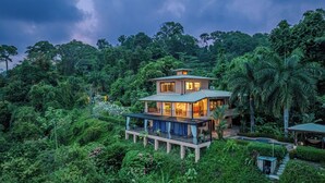 A two-story house surrounded by lush greenery, with a balcony on the upper floor.