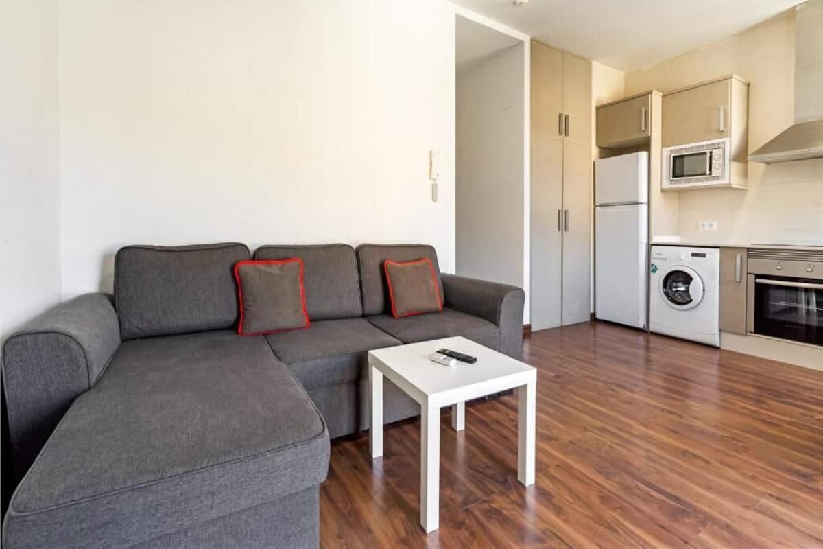 Een moderne woonkamer met een grijze modulaire bank, een witte salontafel en een keukenhoek met een wasmachine en magnetron.