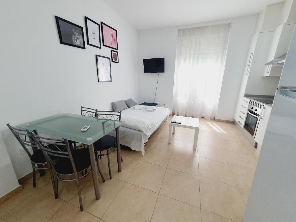 Habitació compacta amb un llit, una taula de dinar de vidre amb cadires, una taula petita blanca, una televisió penjada a la paret i una zona de cuina amb armaris i un frigorífic.
