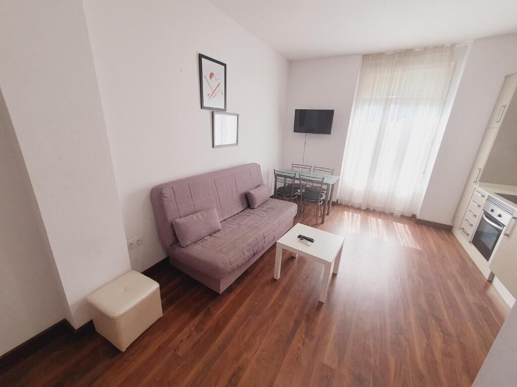 A small living room with a sofa, a coffee table, a TV mounted on the wall, a dining table with chairs, and a kitchen area with cabinets and a stove.