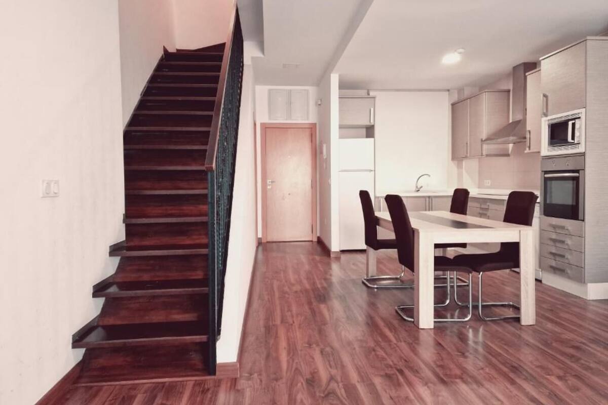 Een moderne keuken met roestvrijstalen apparatuur, een eetgedeelte met een houten tafel en stoelen, en een trap met donkere houten treden.