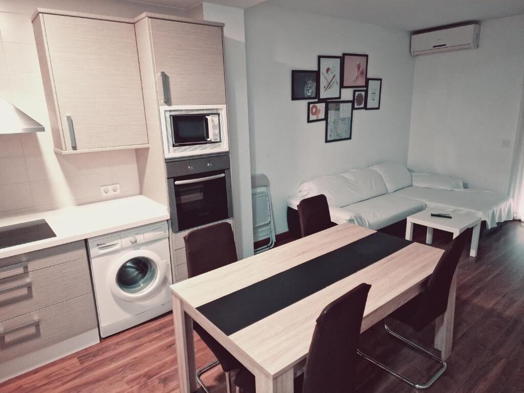 Cocina compacta con microondas, horno y lavadora. Hay una mesa de comedor con sillas y una cama al fondo.