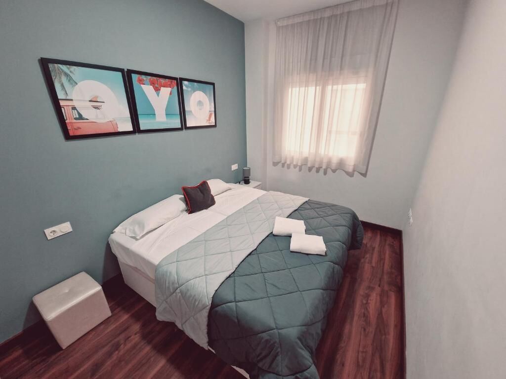 A bedroom with a bed, a nightstand, a window with sheer curtains, and wall art.