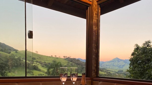 A hot tub with two glasses and a bottle, a wooden tray, and a view of a green landscape through a large window. - Cabana I nas Montanhas de Gonçalves (Gonçalves)