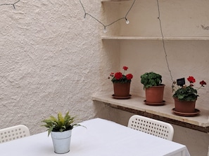 Uma mesa com uma planta em vaso, uma cadeira branca e uma estante com três vasos de terracota contendo flores vermelhas e folhagem verde.