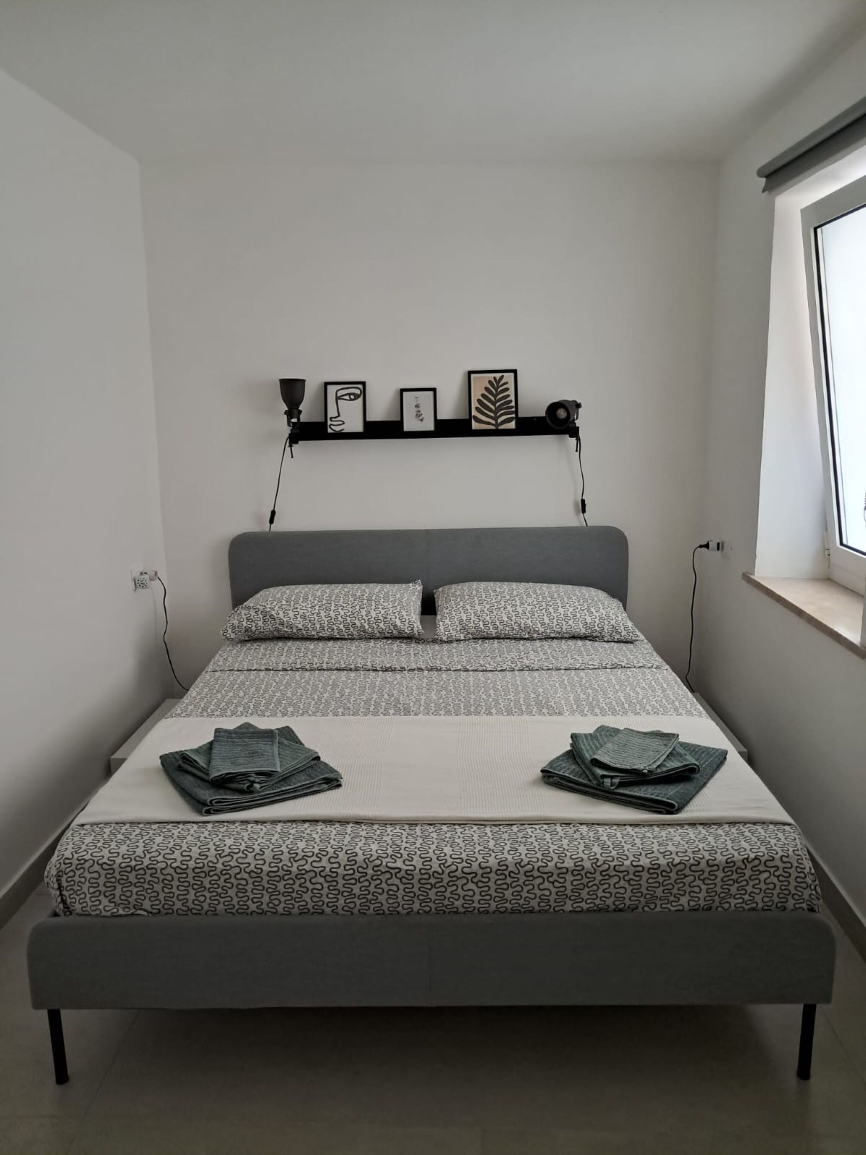 Een moderne slaapkamer met een grijs bed, beddengoed met een patroon, en een plank met ingelijste kunstwerken boven het bed.