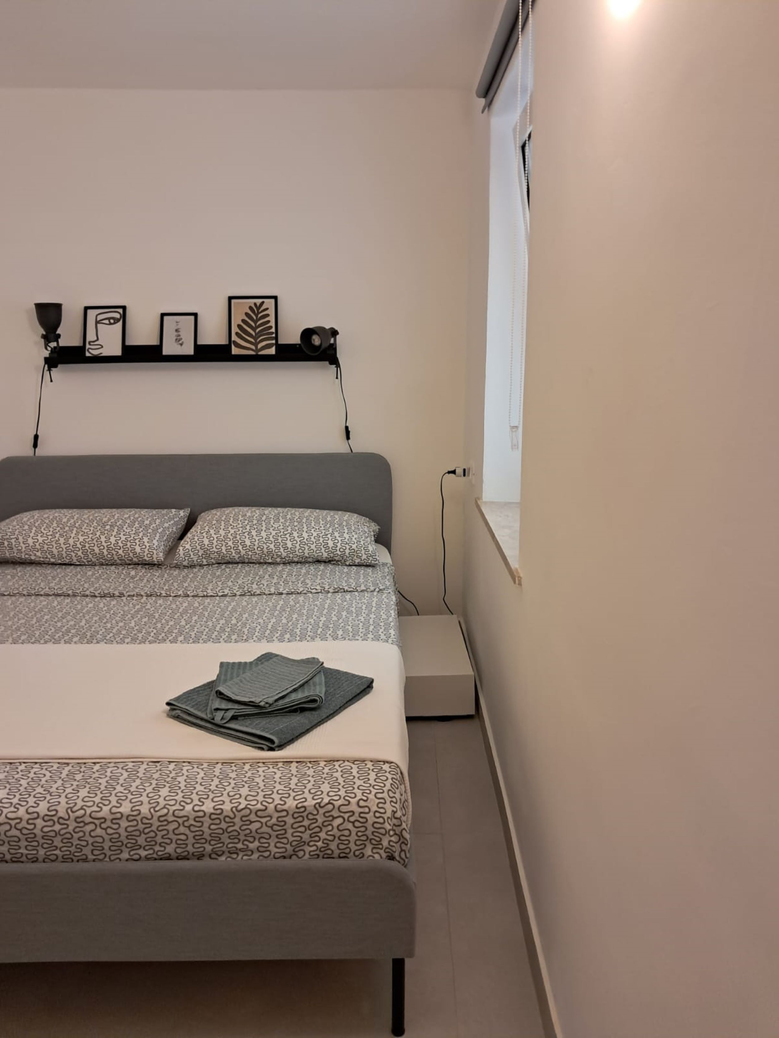 A neatly made bed with patterned bedding, a bedside table with a folded towel, and a wall-mounted shelf with framed artwork.