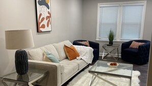 En moderne stue med sofa, to lænestole, et sofabord i glas, en lampe og et billede på væggen.