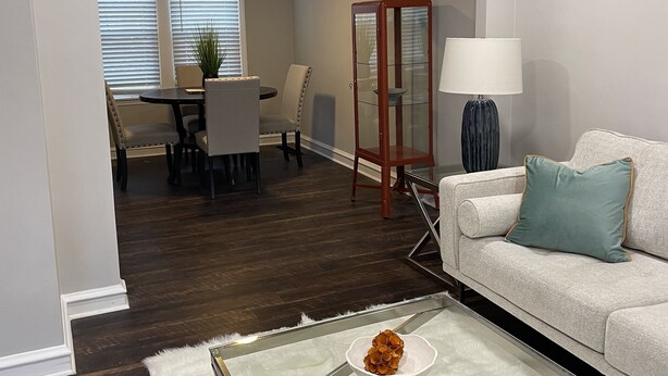 En moderne stue med et glasbord, en hvid sofa med en teal farvet pude, et spiseområde med bord og stole, samt et dekorativt skab.