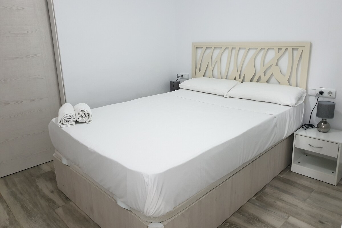 A neatly made bed with white linens, a wooden headboard, a bedside table with a lamp, and a small drawer unit.