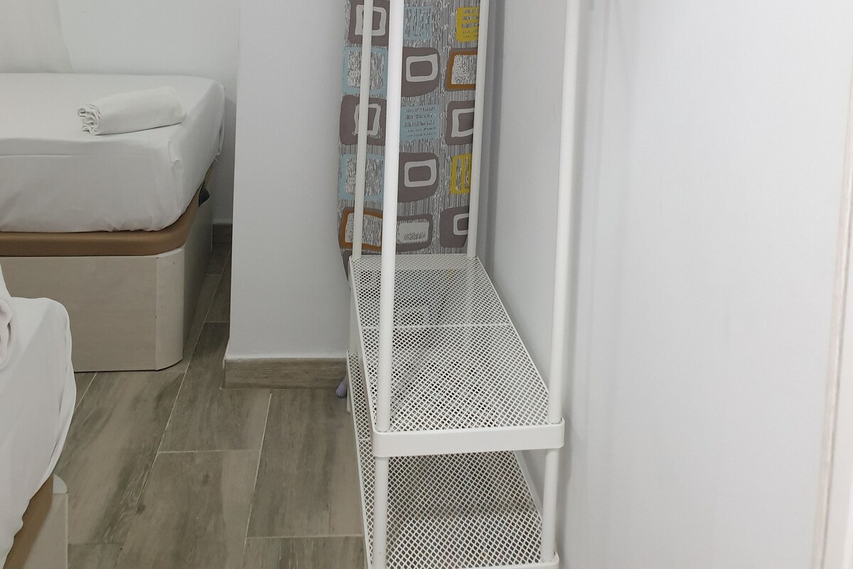 A white bunk bed with a metal frame, a small white shelf, and a patterned curtain in a room with a wooden floor and white walls.