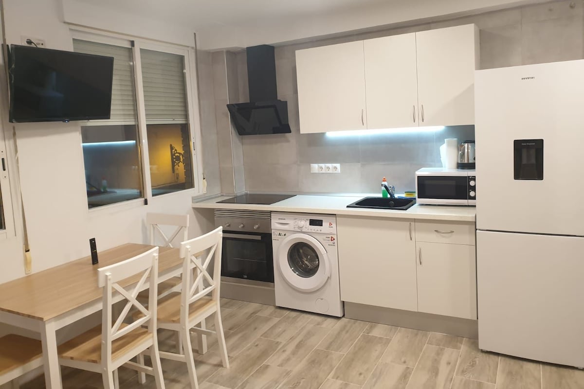 A compact kitchen with a washing machine, microwave, and a dining area with a table and chairs.