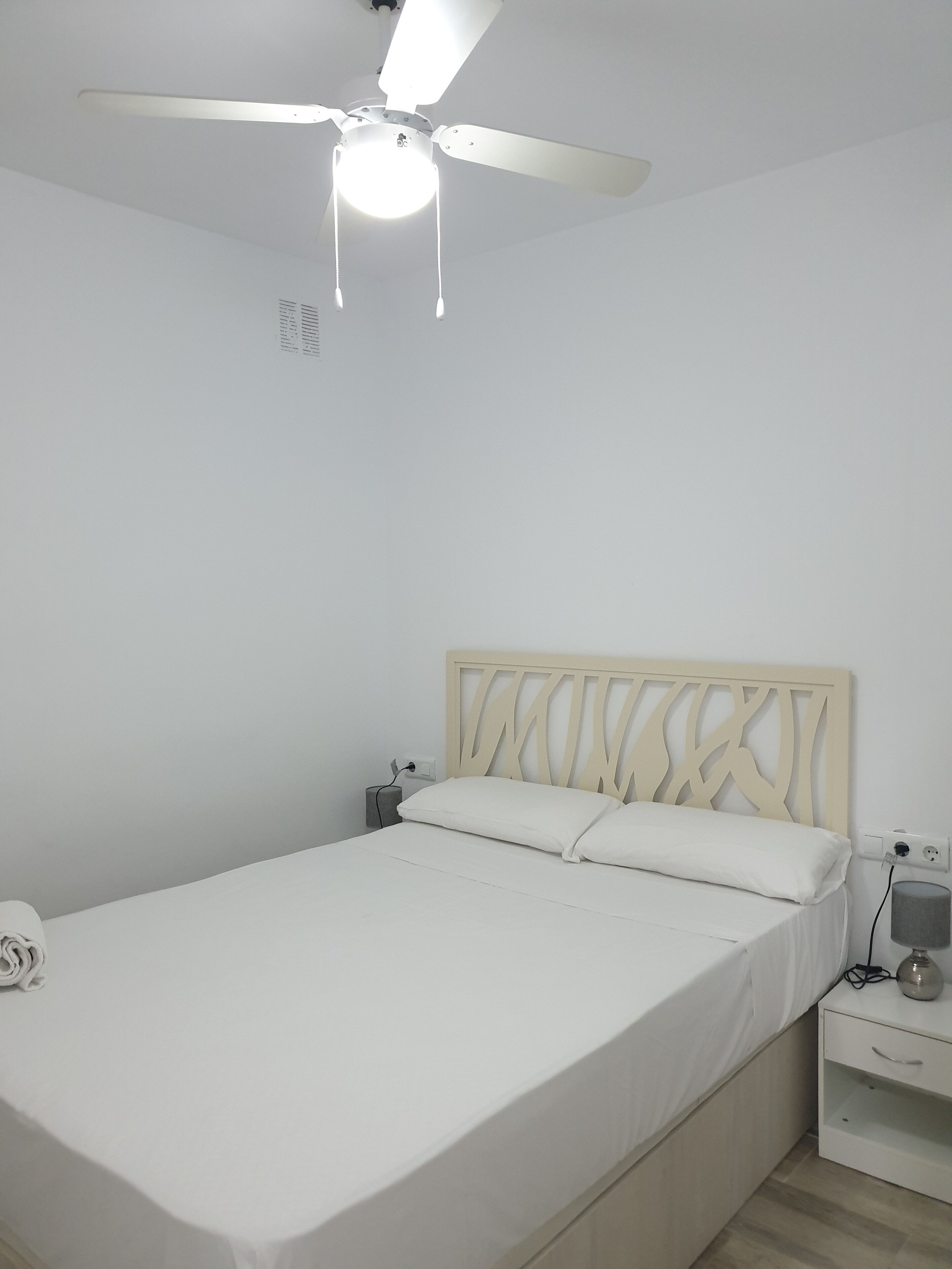 A bedroom with a bed, a nightstand, a lamp, a ceiling fan, and a power outlet.