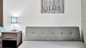 A modern bedroom with a grey sofa, a bedside table with a lamp, and a wall-mounted artwork that reads "Life is better at the lake".
