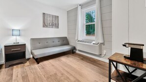 A modern living room with a grey sofa, a small table with a coffee maker, a side table with a lamp, and a window with curtains.