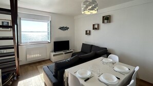 A modern living room with a sofa, a dining table set for two, a television, and a window with a view.