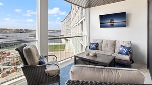 Een balkon met rieten meubels, een bank met kussens en een schilderij van een boom aan de muur.