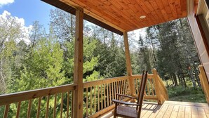 A wooden deck with a ceiling fan, a rocking chair, and a view of a forest. - Riverfront Cabin: The Cozy Southern Brook Trout Lodge  (Luzene)