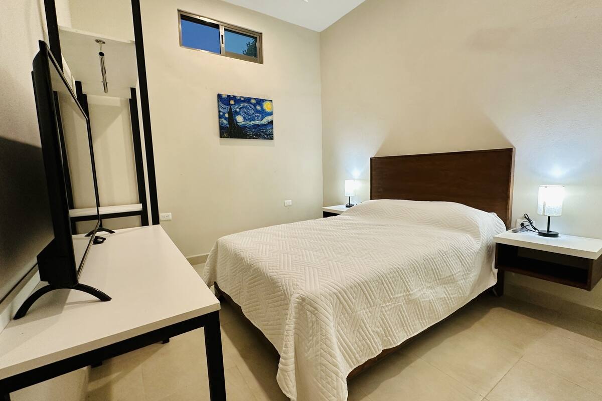 Ein Hotelzimmer mit einem Bett, einem Fernseher, einem Nachttisch, einer Lampe und einem Bild an der Wand.