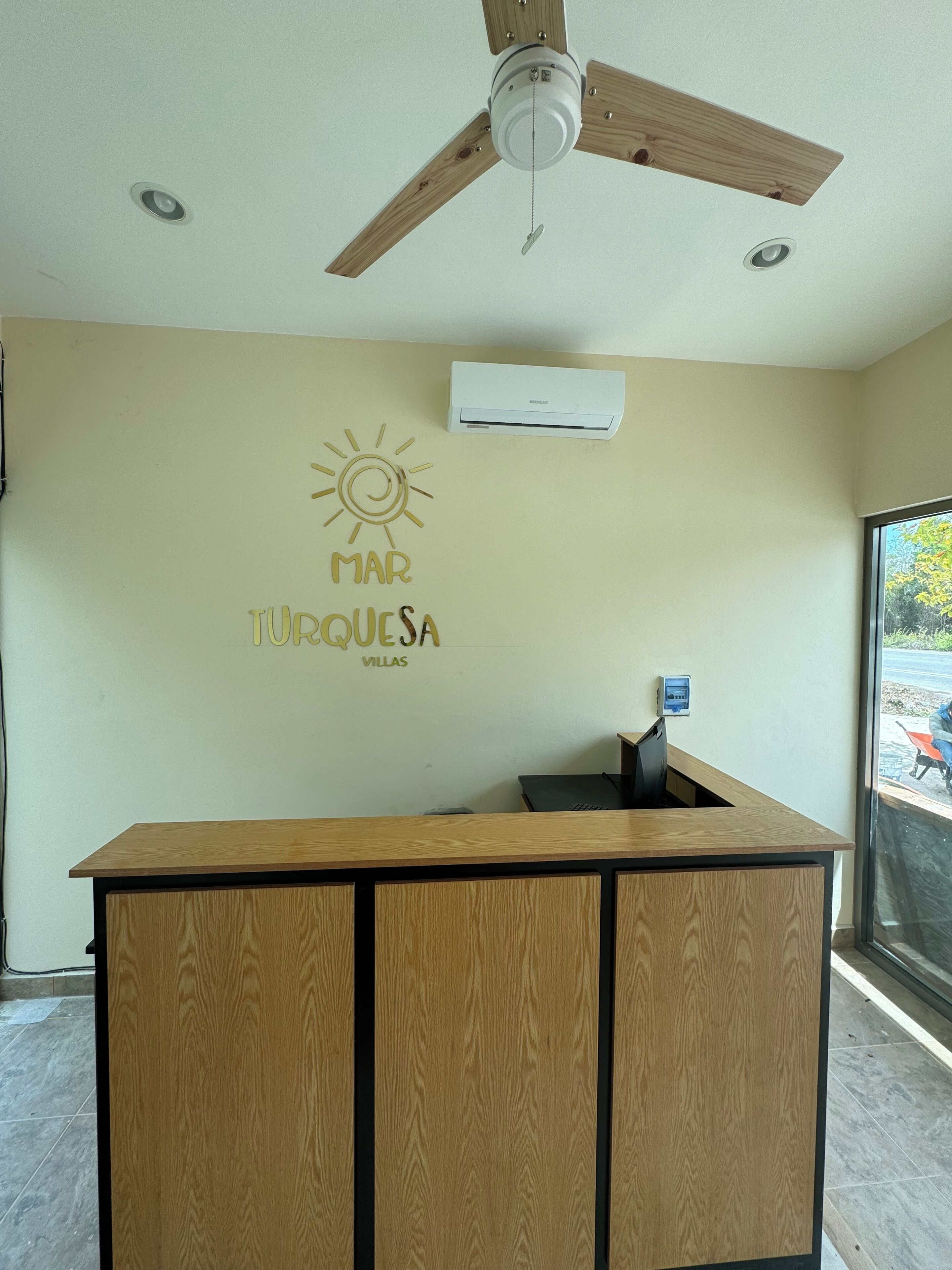 A reception desk with a ceiling fan, an air conditioning unit, and a wall with a sun and text.