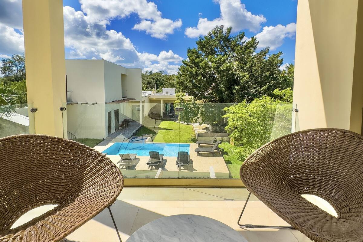 Ein Balkon mit zwei Korbstühlen, einem Marmortisch und Blick auf ein Schwimmbecken und Grünflächen.