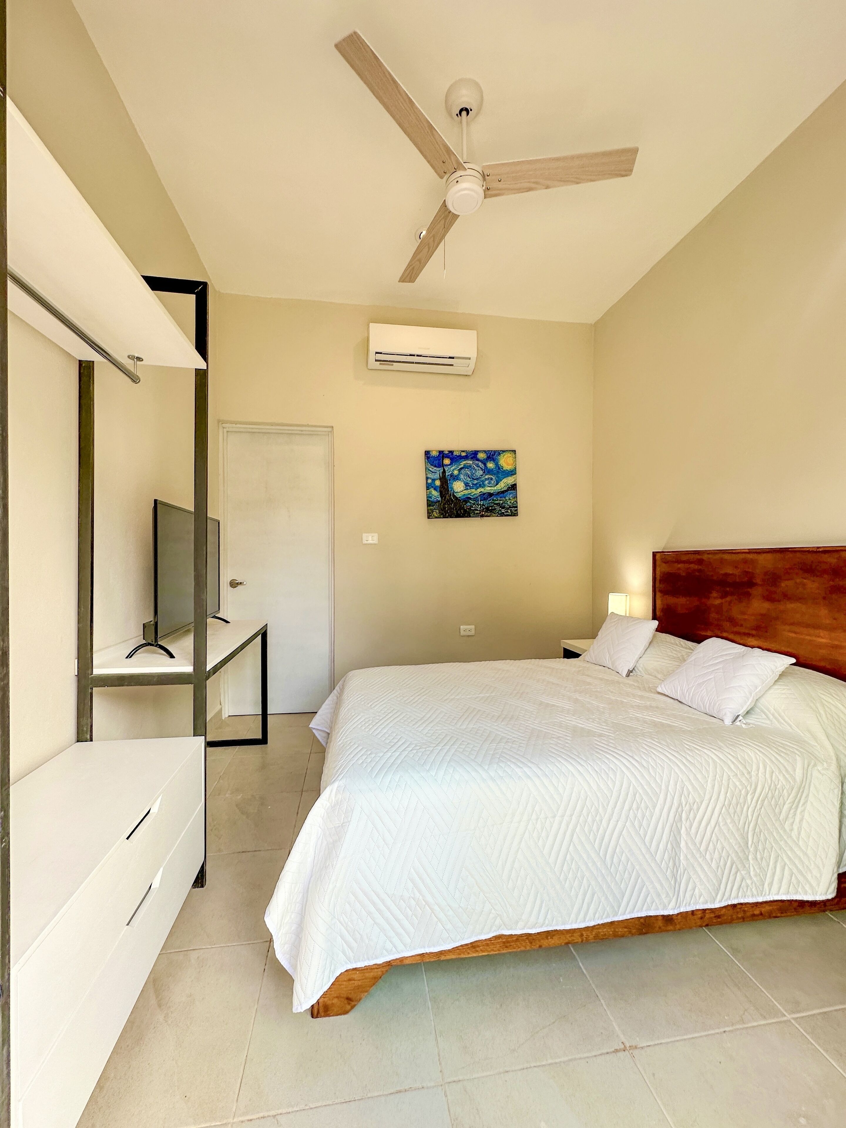 Un dormitorio con cama, ventilador de techo, equipo de aire acondicionado, un cuadro y un espejo.