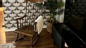 Ein Wohnzimmer mit gemustertem Wandpapier, einem Holzschaukelstuhl, einem Blumentopf und einer schwarzen Unterhaltungsecke.