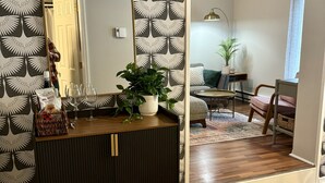 Ein Zimmer mit einem dunklen Holzschrank, einem großen Spiegel und einem Blumentopf. Eine Tür führt in einen weiteren Raum, und ein kleiner Tisch mit einer Lampe und einer Pflanze steht dort.